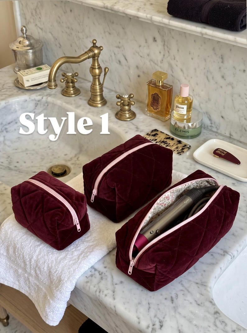 May include: Three burgundy velvet cosmetic bags with a quilted diamond pattern and light pink zippers. The bags vary in size, with one open bag containing a grey hair styling tool. The text "Style 1" is visible in white on the left side.