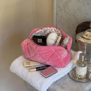 May include: A pink quilted velvet cosmetic bag with a floral interior lining, containing a Givenchy compact, a Chanel hand cream, and two metallic tubes. Outside the bag are a cream hair clip, a black eyeliner, and a pink Dior blush compact.