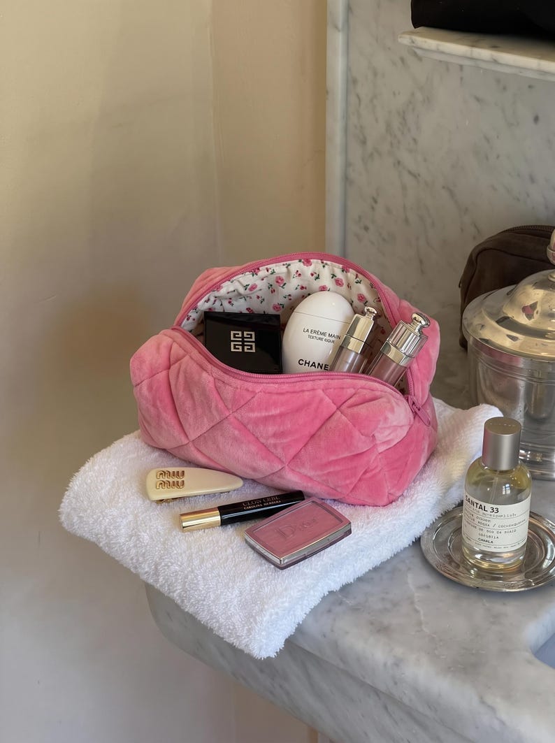 May include: A pink quilted velvet cosmetic bag with a floral interior lining, containing a Givenchy compact, a Chanel hand cream, and two metallic tubes. Outside the bag are a cream hair clip, a black eyeliner, and a pink Dior blush compact.