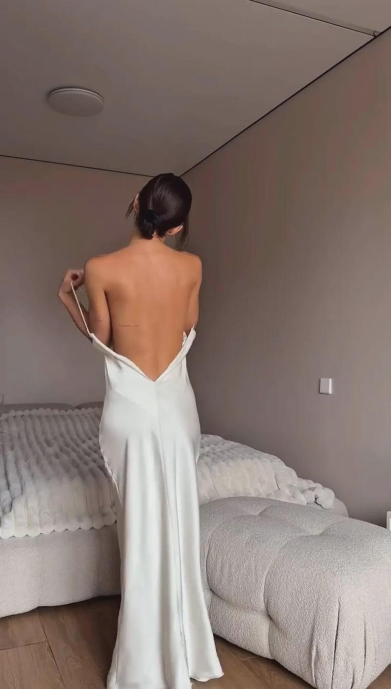 May include: A white satin maxi dress with a low back. The dress has thin straps and a flowing skirt. The model is standing in a bedroom with a bed and a neutral-colored wall.