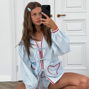 Puede incluir: Conjunto de pijama de cuadros vichy azul claro con ribete rojo. El conjunto incluye una parte superior de manga larga con cierre de lazo y bolsillo en forma de coraz&oacute;n, y pantalones cortos a juego. Los pu&ntilde;os y los bordes est&aacute;n ribeteados en rojo.