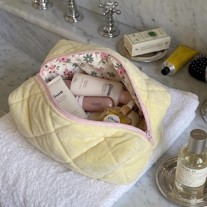 May include: A pale yellow quilted cosmetic bag with a light pink zipper and a floral interior lining. The bag is filled with various skincare and beauty products, including bottles labeled Gisou and Kiko, visible through the open top zipper.