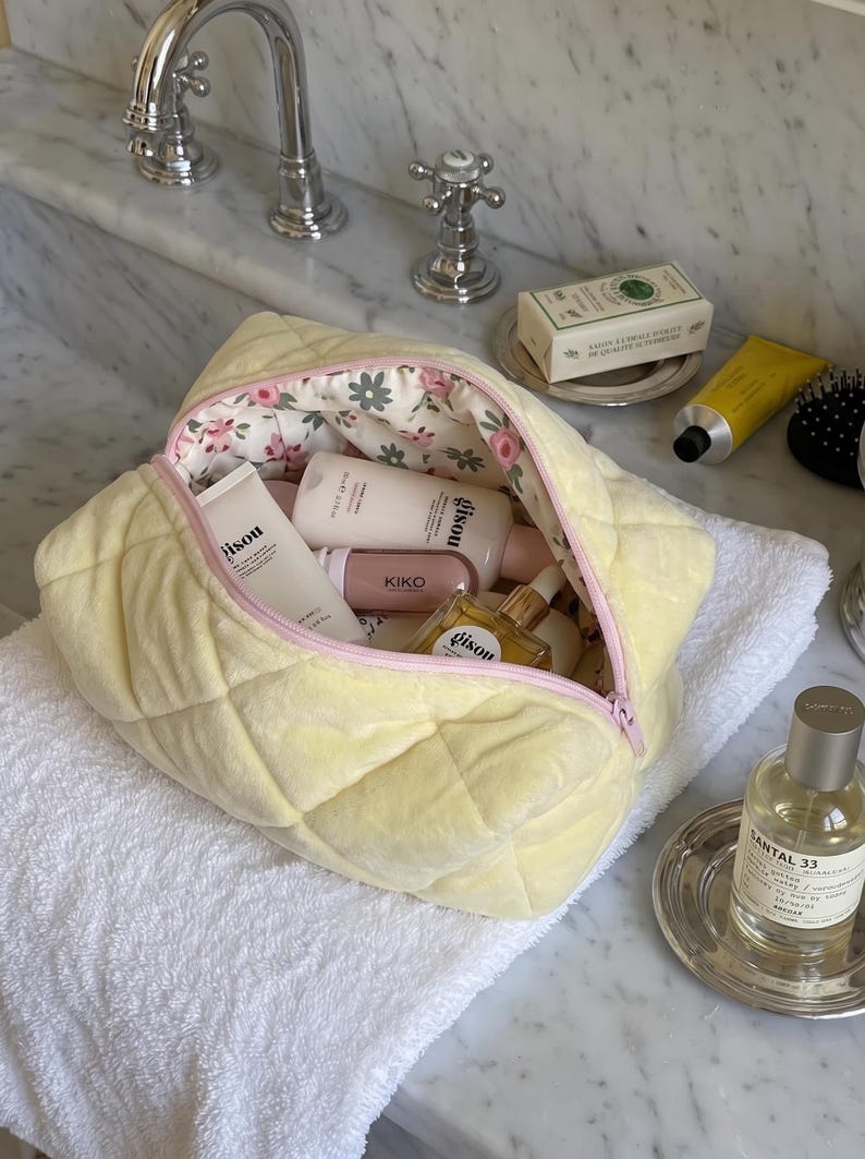 May include: A pale yellow quilted cosmetic bag with a light pink zipper and a floral interior lining. The bag is filled with various skincare and beauty products, including bottles labeled Gisou and Kiko, visible through the open top zipper.