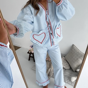Puede incluir: Conjunto de pijama de cuadros vichy azul claro y blanco con ribete rojo. La parte superior tiene bolsillos en forma de coraz&oacute;n y un dobladillo con volantes. El conjunto es visible en un espejo.