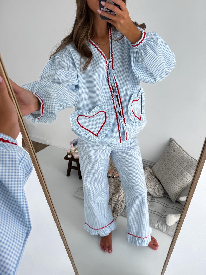 Puede incluir: Conjunto de pijama de cuadros vichy azul claro y blanco con ribete rojo. La parte superior tiene bolsillos en forma de coraz&oacute;n y un dobladillo con volantes. El conjunto es visible en un espejo.