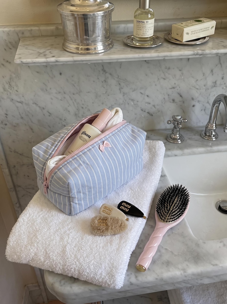 May include: A light blue and white striped fabric wash bag with a pink zip and a small pink bow detail. The bag contains a Gisou bottle and white fabric. Nearby are two Miu Miu hair clips, a fuzzy hair accessory, and a pink hairbrush.