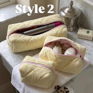 May include: Three pale yellow quilted pouches with soft fabric exteriors and light pink zippers. The largest pouch contains a metallic hair styling tool, the medium pouch holds various skincare bottles, and the smallest pouch is empty.