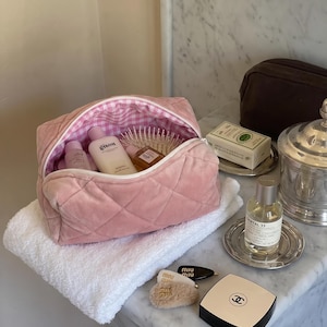 May include: A pink quilted velvet cosmetic bag with a white and pink gingham interior lining. The bag is open and contains several skincare bottles, including a Gisou bottle, and a hairbrush. Nearby are a Chanel compact and Miu Miu hair clips.