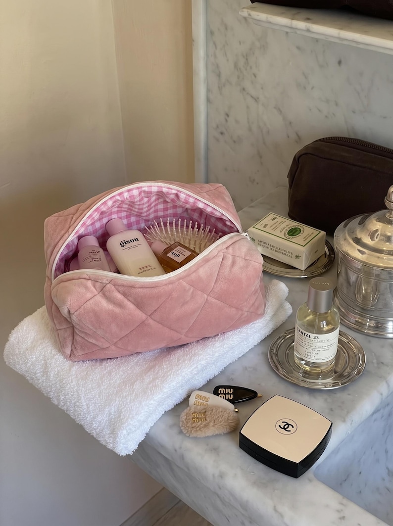 May include: A pink quilted velvet cosmetic bag with a white and pink gingham interior lining. The bag is open and contains several skincare bottles, including a Gisou bottle, and a hairbrush. Nearby are a Chanel compact and Miu Miu hair clips.