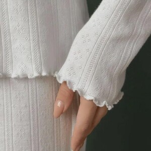 May include: A white long-sleeved top featuring a vertical ribbed texture with a subtle diamond pattern throughout. The garment is finished with a delicate lettuce-edge hem at the sleeve cuff, providing a soft, wavy detail to the fabric edge.