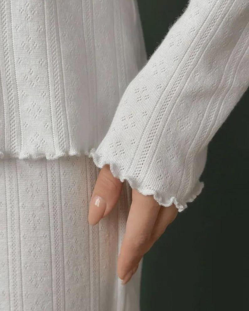 May include: A white long-sleeved top featuring a vertical ribbed texture with a subtle diamond pattern throughout. The garment is finished with a delicate lettuce-edge hem at the sleeve cuff, providing a soft, wavy detail to the fabric edge.