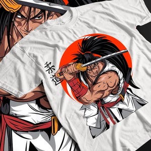 May include: White t-shirt featuring a detailed illustration of a samurai warrior holding a katana sword against a red sun. The design includes Japanese text and is printed on a white cotton t-shirt.