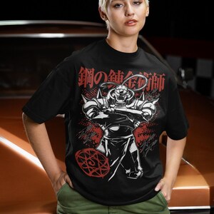 May include: Black t-shirt featuring a graphic of a character from the anime series Fullmetal Alchemist. The design includes the Japanese text "鋼の錬金術師" in red and white. The character is depicted in a dynamic pose with arms crossed.