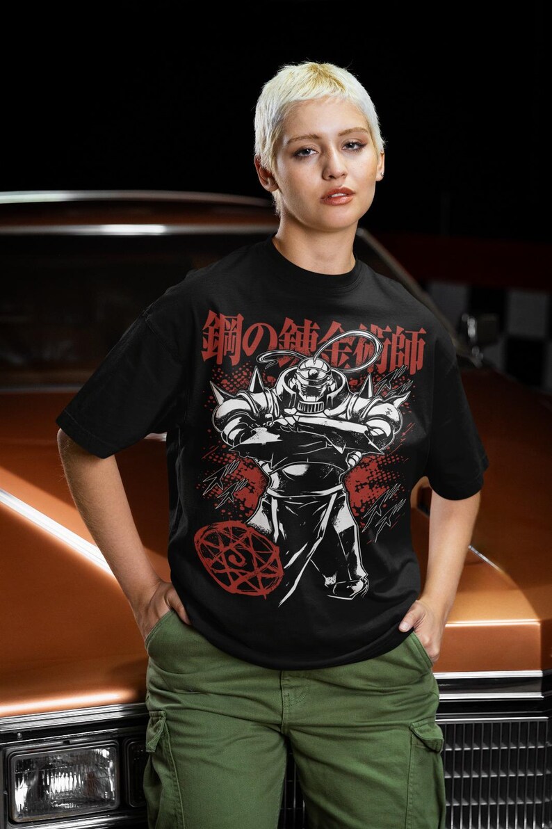 May include: Black t-shirt featuring a graphic of a character from the anime series Fullmetal Alchemist. The design includes the Japanese text "鋼の錬金術師" in red and white. The character is depicted in a dynamic pose with arms crossed.