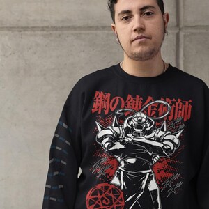 May include: Black sweatshirt featuring a graphic of a character from the anime series Fullmetal Alchemist. The design includes Japanese text and a red alchemic circle. The sleeve has a pattern of blue and gray lines.