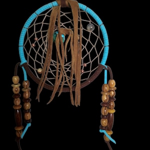 May include: A handcrafted dreamcatcher with a turquoise-colored hoop and a web of white string. Brown leather strips hang from the center, adorned with beads in various shades of brown and tan. A wooden bead tops the dreamcatcher.