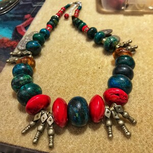 May include: A handcrafted necklace featuring a variety of beads in shades of red, green, and blue. The necklace includes large, round, and oval beads, along with silver-toned metal accents. The necklace is displayed on a tan surface.