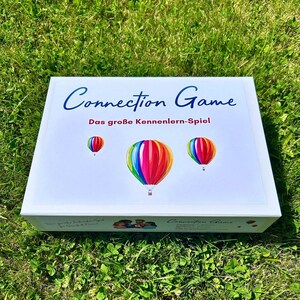 Könnte beinhalten: Eine weiße Spielbox mit dem Titel "Connection Game" in blauer Schrift. Die Box zeigt drei bunte Heißluftballons. Der Text "Das große Kennenlern-Spiel" ist ebenfalls vorhanden. Die Box liegt auf grünem Gras.