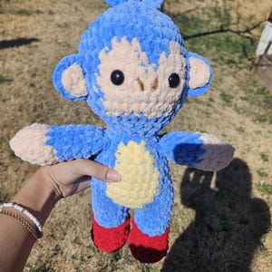 May include: A handmade blue crochet monkey plushie with a light tan face, ears, and belly. The monkey has black safety eyes, red feet, and is held up by a person. The plushie is a soft, cuddly toy.
