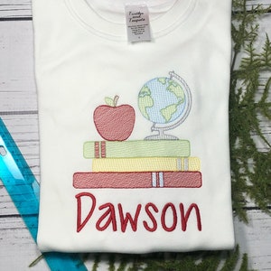 May include: White t-shirt with red embroidery of a stack of books, an apple, and a globe. The name "Dawson" is embroidered below the books.