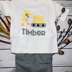 May include: A white short-sleeved shirt with a yellow construction vehicle holding candy corn. The vehicle has a spider web and a bat on the arm. The shirt has the name "Timber" embroidered on it. The shirt is paired with black and white checkered pants.