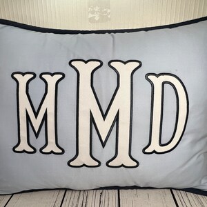 May include: A light blue rectangular pillow with a navy blue border. The pillow features the embroidered letters "MMD" in white with a black outline. The pillow is made of a soft fabric.
