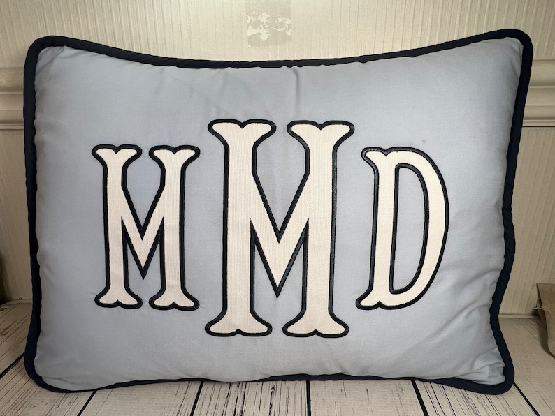 May include: A light blue rectangular pillow with a navy blue border. The pillow features the embroidered letters "MMD" in white with a black outline. The pillow is made of a soft fabric.
