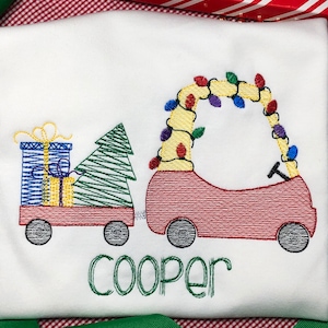 May include: A white t-shirt with an embroidered design of a red toy car pulling a wagon with a Christmas tree and presents. The car is decorated with colorful lights. The name "Cooper" is embroidered below the design.