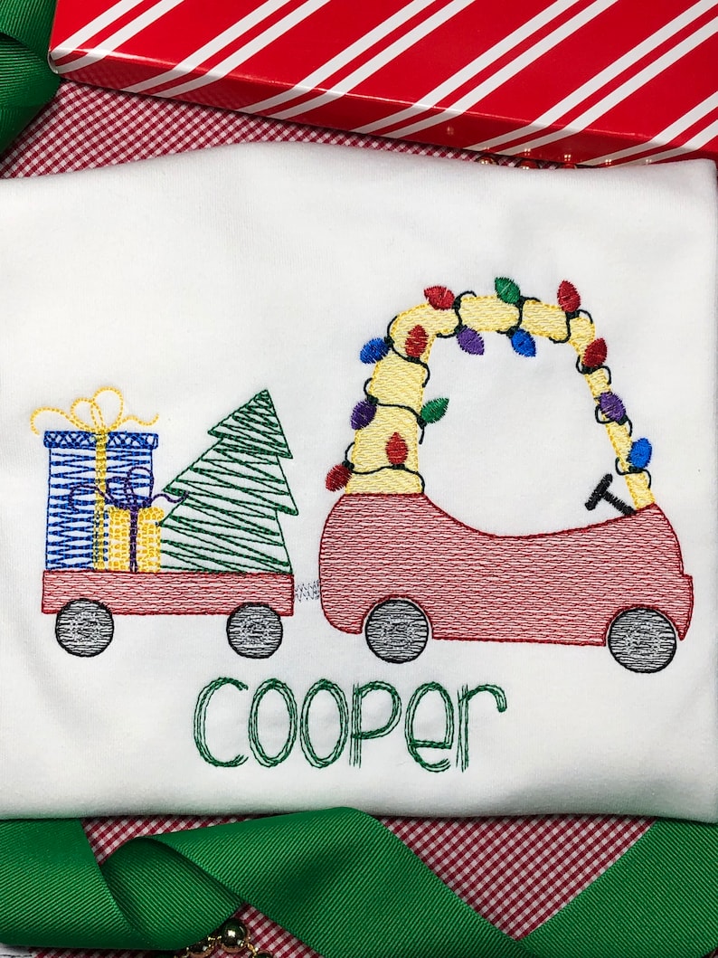 May include: A white t-shirt with an embroidered design of a red toy car pulling a wagon with a Christmas tree and presents. The car is decorated with colorful lights. The name "Cooper" is embroidered below the design.