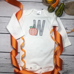 May include: White long-sleeve onesie with a blue embroidered letter W and a pumpkin with a red and white checkered pattern. The onesie has a small tag that says "Twirly and Twigs".