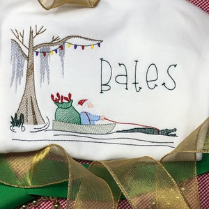 Pode incluir: Desenho bordado de um Papai Noel em um barco com um saco de presentes, uma &aacute;rvore de cipreste com musgo espanhol e luzes de Natal. O texto "Bates" est&aacute; bordado acima da cena.