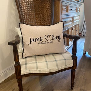 May include: A vintage wooden armchair with a woven back and plaid seat cushion. A rectangular pillow with the names "Jamie & Trey" and the date "February 16, 2019" is on the seat. The pillow has a black border.