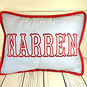 May include: A rectangular pillow with the name "WARREN" embroidered in red, outlined in white, on a light blue background. The pillow has a red border.