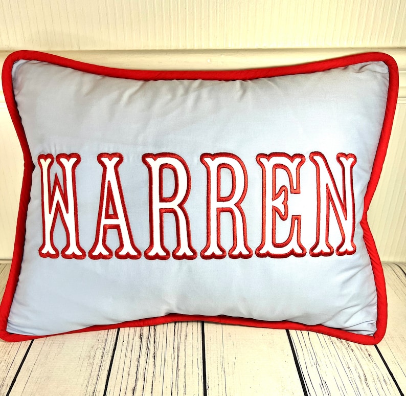 May include: A rectangular pillow with the name "WARREN" embroidered in red, outlined in white, on a light blue background. The pillow has a red border.