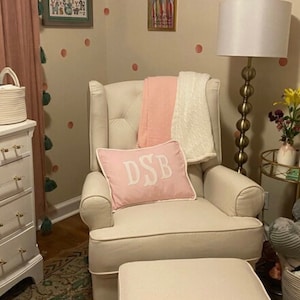 May include: A white upholstered rocking chair with a pink throw blanket and a pink pillow with the monogram "DSB" in white. The chair has a matching white ottoman. The chair is in a room with a pink and green patterned rug and a lamp with a white shade.