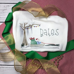 Pode incluir: Camiseta branca bordada com tema natalino com uma imagem de desenho animado do Papai Noel em um barco sob uma &aacute;rvore com luzes. O texto "Bates" est&aacute; bordado na camiseta.