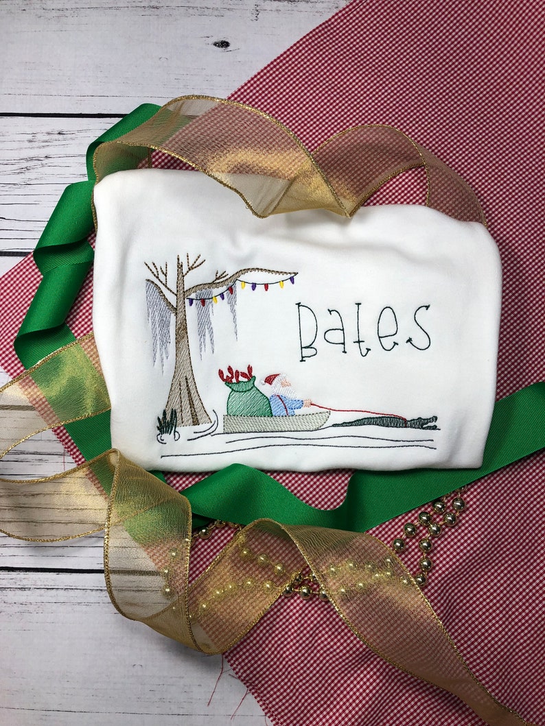 Pode incluir: Camiseta branca bordada com tema natalino com uma imagem de desenho animado do Papai Noel em um barco sob uma &aacute;rvore com luzes. O texto "Bates" est&aacute; bordado na camiseta.