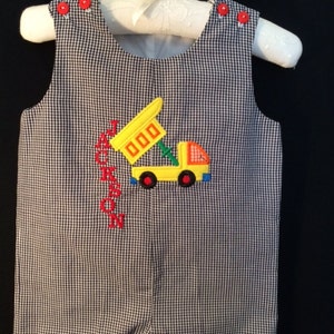 May include: A black and white gingham baby onesie with red buttons and an embroidered yellow construction truck with the name "Jackson" in red.