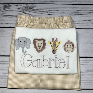 May include: A two-piece baby outfit featuring a white top with embroidered animal faces and the name "Gabriel" in brown thread. The shorts are tan and white gingham. The top has an elephant, lion, giraffe, and monkey.