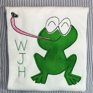 May include: A white t-shirt with a green frog applique. The frog has a pink tongue sticking out and is catching a black butterfly. The letters WJH are embroidered on the shirt.