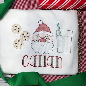 May include: White long-sleeve shirt with an embroidered design of Santa Claus, cookies, a glass of milk, and a straw. The name "Caillan" is embroidered below the design in red and green thread.