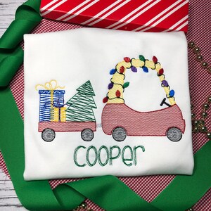 May include: A white Christmas-themed embroidered shirt with a red car carrying a Christmas tree and presents. The car is decorated with colorful lights. The name "Cooper" is embroidered below the car.