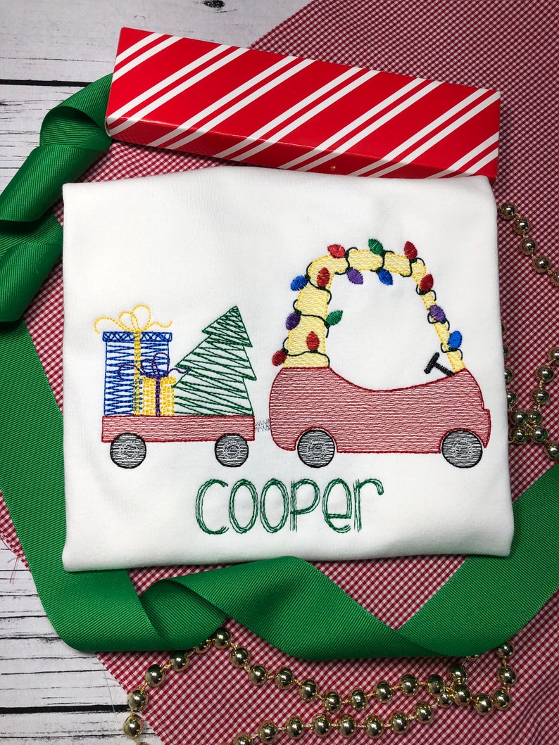 May include: A white Christmas-themed embroidered shirt with a red car carrying a Christmas tree and presents. The car is decorated with colorful lights. The name "Cooper" is embroidered below the car.