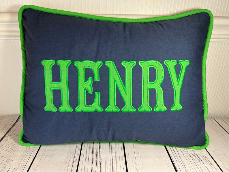 May include: Navy blue rectangular pillow with the name "HENRY" embroidered in bright green. The pillow has a green border, creating a contrasting design. The pillow is placed on a white wooden surface.