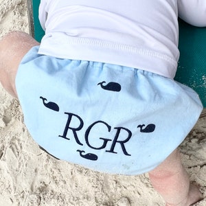 May include: A light blue baby bloomer with a white waistband and embroidered whales. The bloomers have the initials "RGR" embroidered in black.