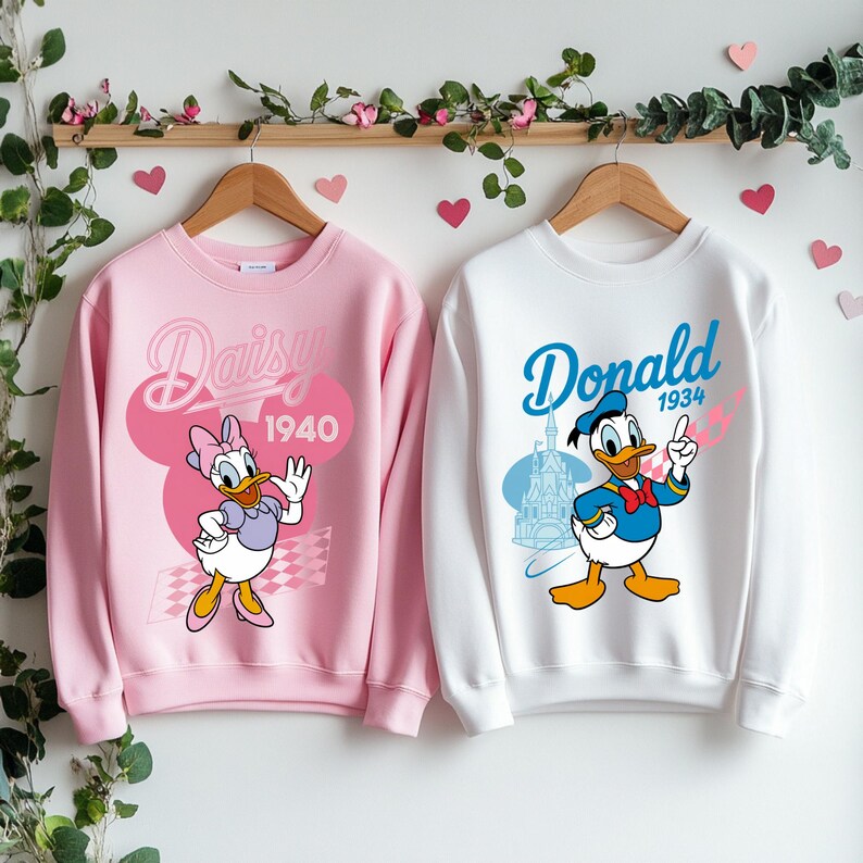 K&ouml;nnte beinhalten: Zwei Sweatshirts, eines rosa und eines wei&szlig;, jeweils mit einer Disney-Figur. Das rosa Sweatshirt hat Daisy Duck mit dem Text "Daisy 1940". Das wei&szlig;e Sweatshirt zeigt Donald Duck mit dem Text "Donald 1934".