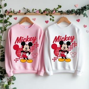 May include: Two sweatshirts, one pink and one white, each featuring a Mickey Mouse graphic. The design includes the word "Mickey" in red script, a cartoon image of Mickey Mouse, and a castle silhouette.