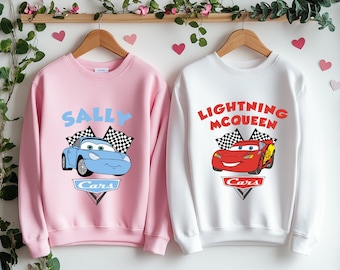 May include: Two pink and white sweatshirts with Cars characters. The pink sweatshirt has a blue car with the text "Sally Cars" and the white sweatshirt has a red car with the text "Lightning McQueen Cars".