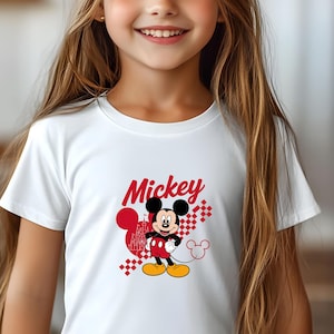 May include: White t-shirt featuring a graphic of Mickey Mouse. The design includes Mickey in red, black, and yellow, with the word "Mickey" in red script. A red heart and castle graphic are also present.