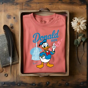 K&ouml;nnte beinhalten: Korallfarbenes T-Shirt mit einer Grafik von Donald Duck, der nach oben zeigt. Das Design beinhaltet das Wort "Donald" in blauer Schrift, das Jahr "1934" und eine Schlossillustration. Das Shirt wird in einem Holztablett pr&auml;sentiert.